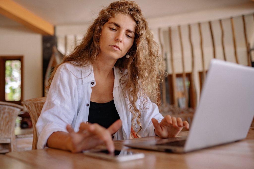 Foto de uma mulher sentanda na frente de um notebook, enquanto mexe também no celular.